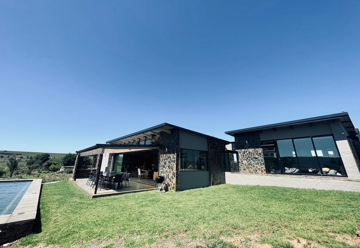 A modern villa with a stone facade is set against a clear blue sky. The structure features expansive glass windows and a spacious outdoor dining area under a covered terrace, leading to a natural saltwater pool and lush green lawn.