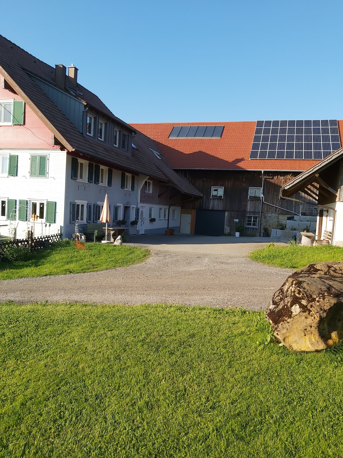 A serene outdoor view features a charming farmhouse structure with green shutters and a red tiled roof, complemented by solar panels. The gravel driveway curves through vibrant green grass, leading toward the main entrance, which is framed by rustic wooden elements.