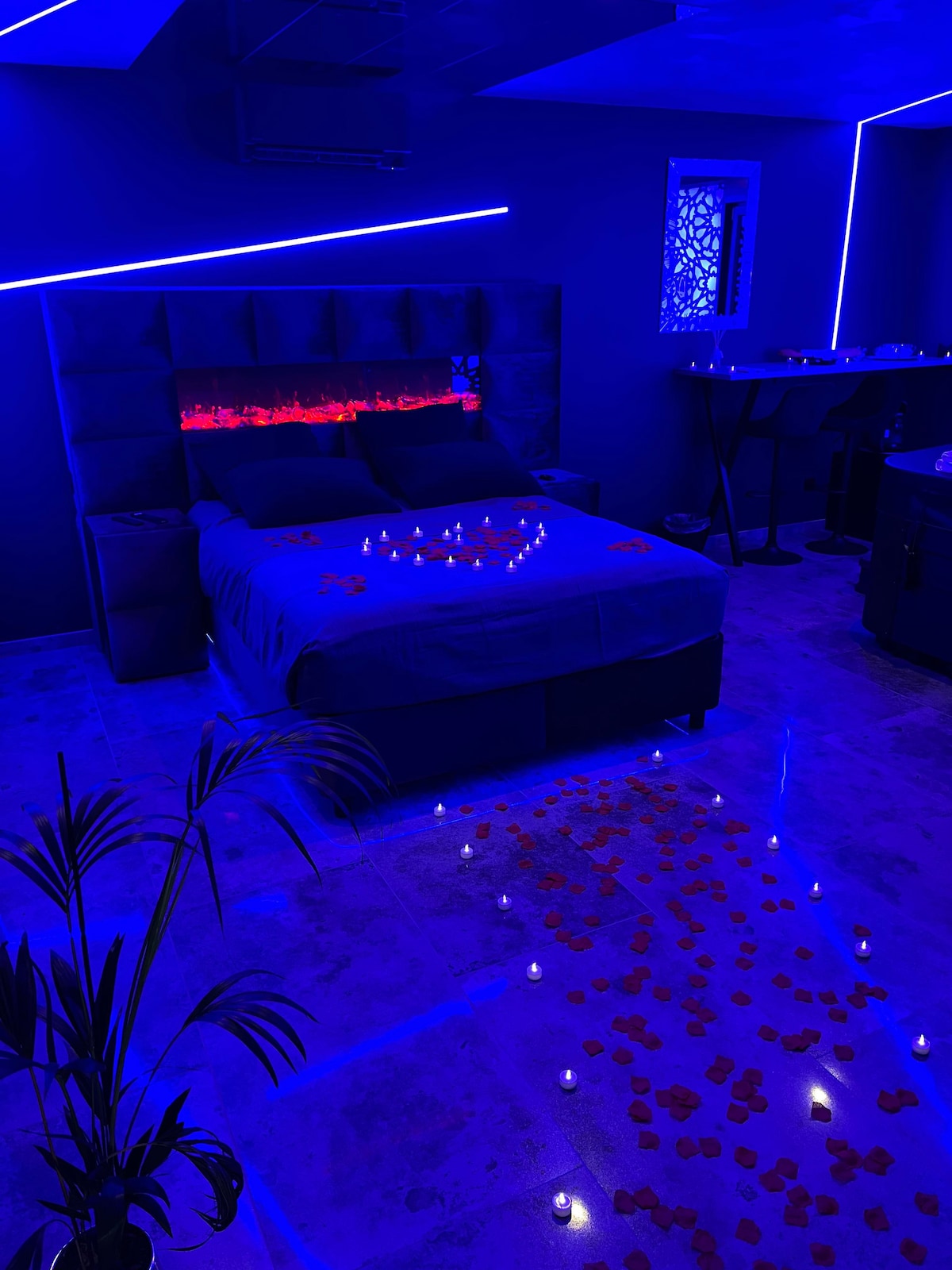 A dimly lit bedroom features a spacious bed adorned with soft, dark bedding. Bright blue LED lights accentuate the room, illuminating scattered rose petals and candles on the floor. A decorative panel with intricate patterns adds character to the wall, enhancing the overall ambiance.