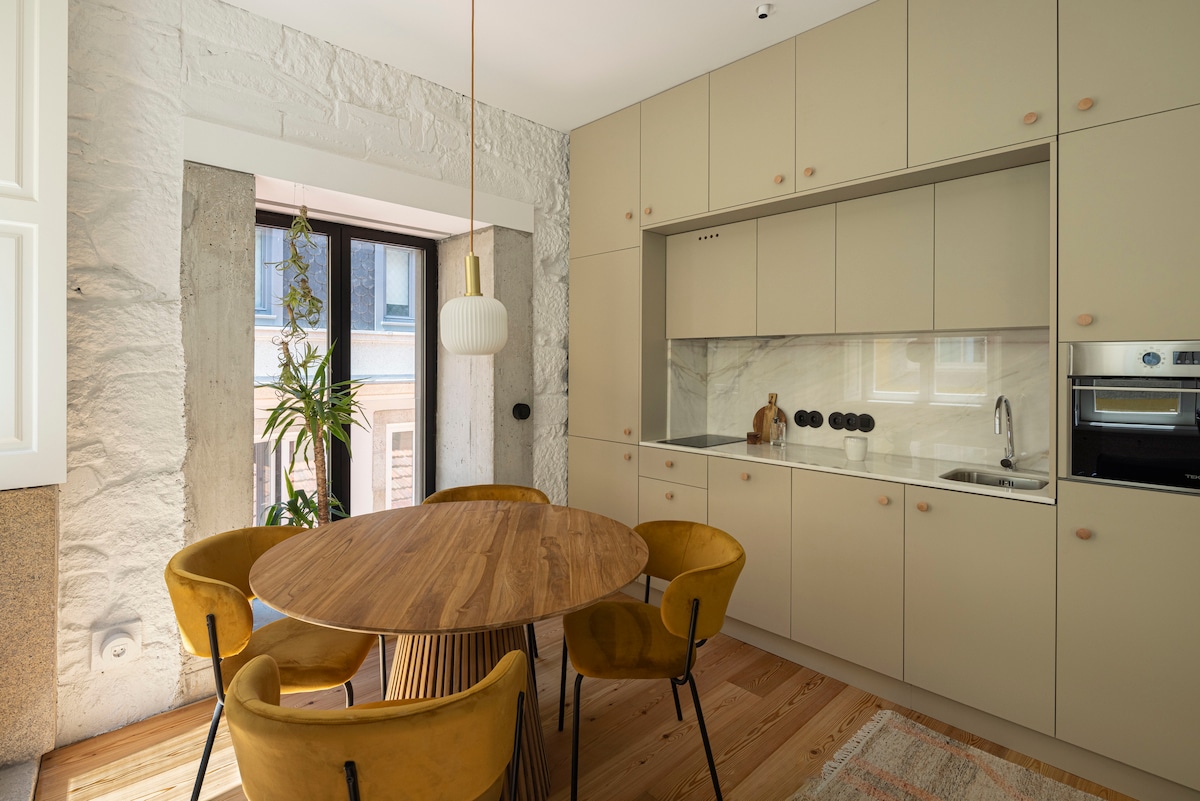 A modern kitchenette is highlighted, featuring a circular wooden dining table surrounded by four mustard-colored chairs. Light walls and stone textures are complemented by a window with a view of greenery. The kitchen is equipped with cabinets and appliances, maintaining a clean and functional aesthetic.