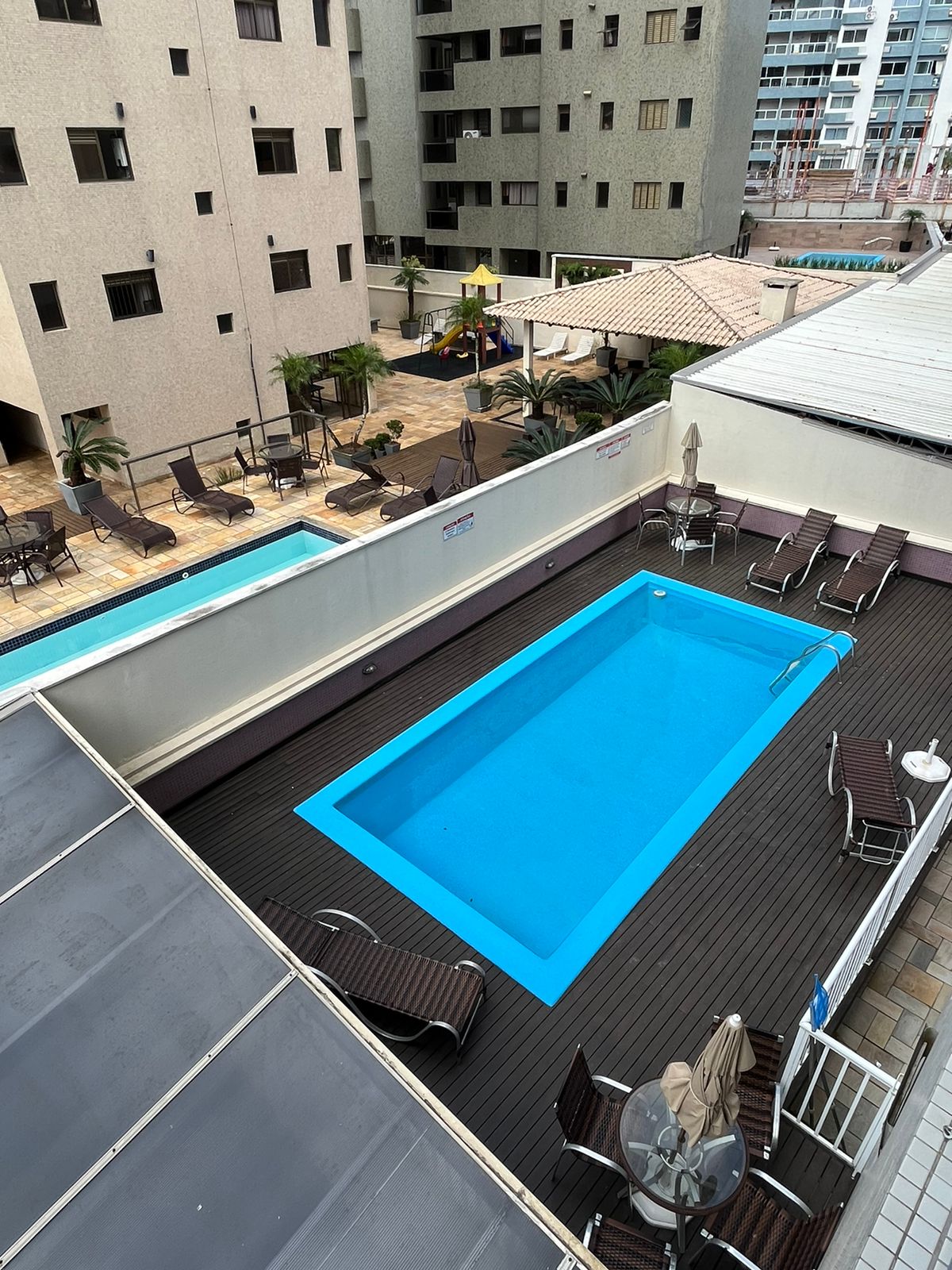A view of a rectangular swimming pool is presented, surrounded by wooden decking. Lounge chairs are arranged along the perimeter, offering space for relaxation. Nearby greenery adds a touch of nature, while surrounding buildings provide structure to the environment.