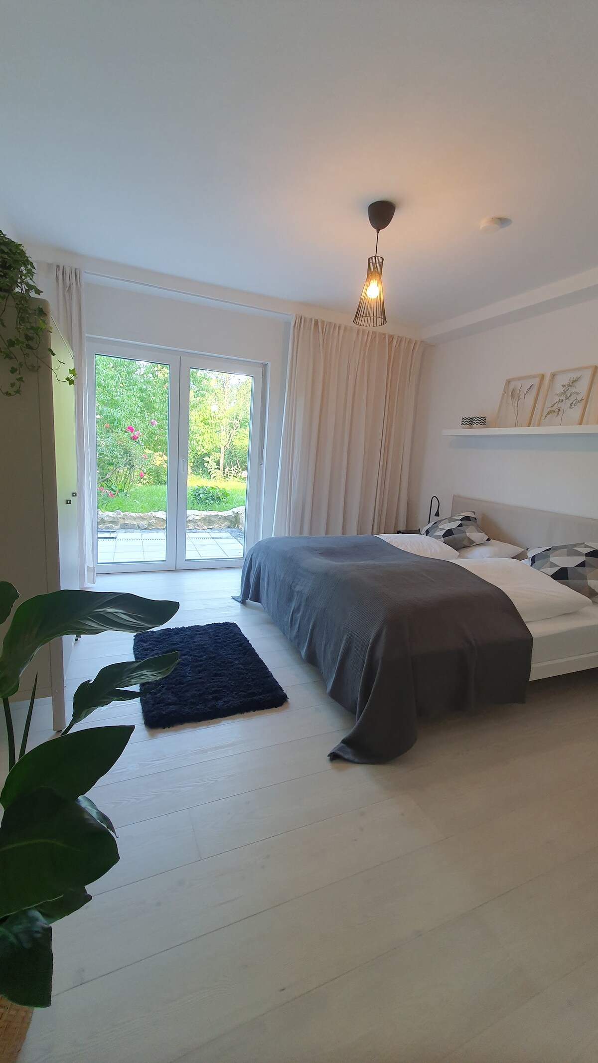 A serene bedroom features a spacious bed draped in neutral bedding. Natural light filters through the glass doors leading to the terrace, surrounded by greenery. A small rug lies on the wooden floor, and a decorative plant adds a touch of nature to the space.
