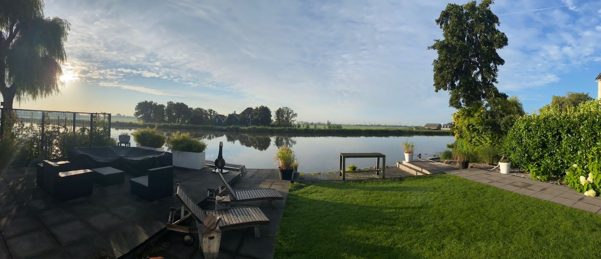 A panoramic view of the waterfront garden showcases a calm river reflecting the morning light. Comfortable seating areas are arranged on a paved patio, surrounded by greenery, while planters and trees add natural beauty to the setting.