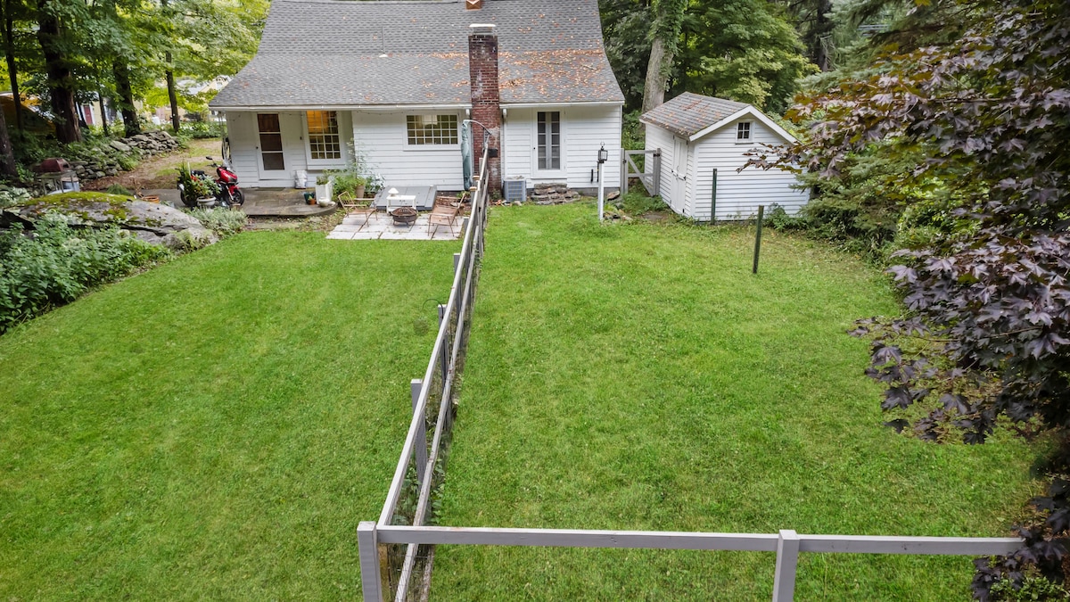 A spacious backyard is enclosed by a white fence, featuring a well-maintained lawn with lush, green grass. The house's exterior is visible in the background, complemented by a small shed. A patio area with seating is accessible from the back door.