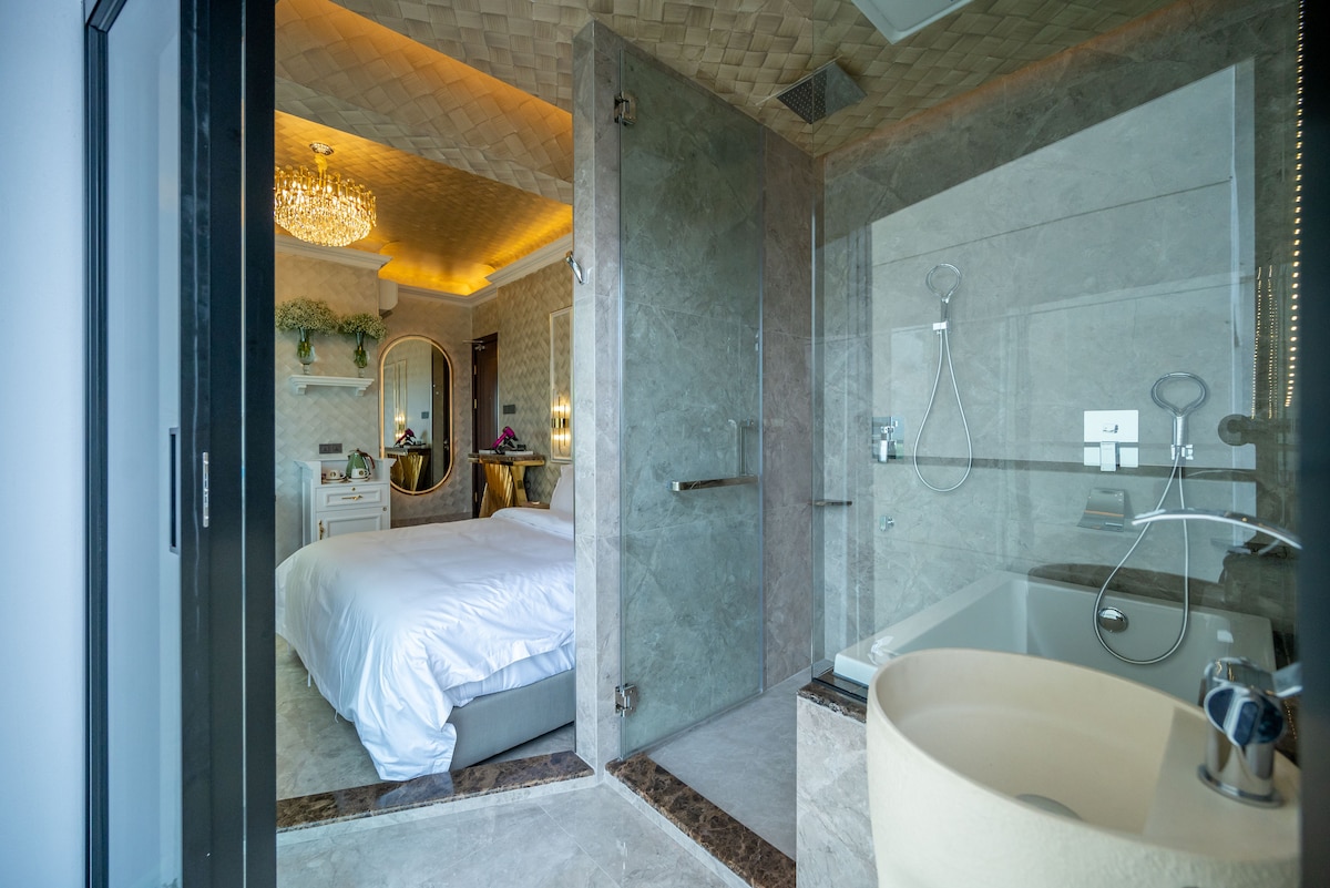A modern bedroom is visible through a glass door, featuring a spacious bed with crisp white linens. The en-suite bathroom displays elegant marble finishes, a glass shower, and a luxurious freestanding tub, illuminated by soft lighting that enhances the tranquil ambiance.