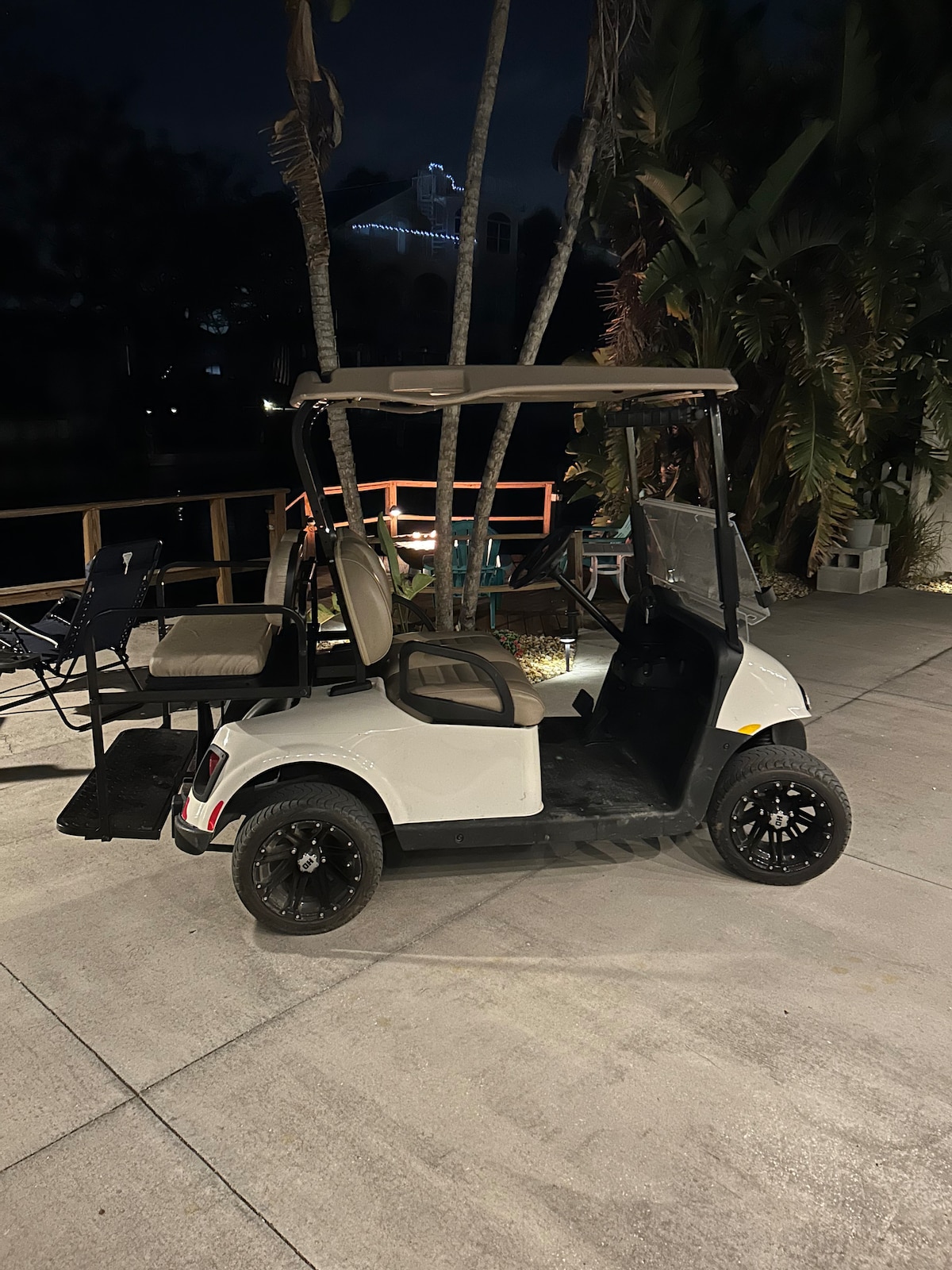 An electric golf cart is positioned on a paved patio, featuring four seats and a spacious storage area at the back. The surrounding area includes tropical plants and faint light from nearby structures, creating a laid-back atmosphere suitable for exploring the local surroundings.