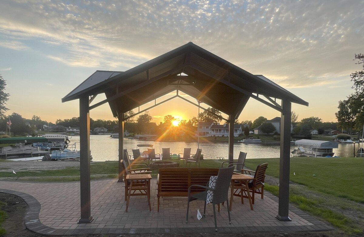 A covered patio with a spacious seating arrangement features wooden chairs and a circular fire pit. The sunset casts a warm glow over the water, highlighting the serene atmosphere. The surrounding area is well-maintained grass, leading towards a calm, inviting body of water.