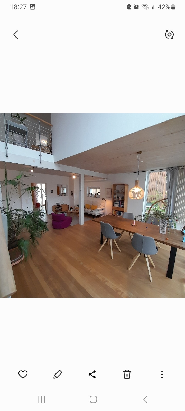 An open-plan living area is showcased with wooden flooring and plenty of natural light. A dining table with grey chairs is positioned at the center, while a colorful armchair adds a pop of color nearby. Large windows provide views of the outdoor space.