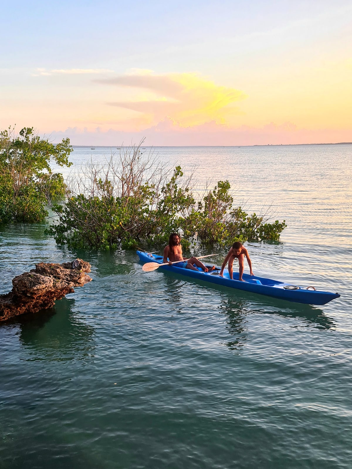 Two individuals are seen in a kayak navigating calm waters, surrounded by greenery and rocky formations. The background features a serene sunset with shades of orange and pink reflecting on the water's surface. The scene captures a peaceful moment in nature.