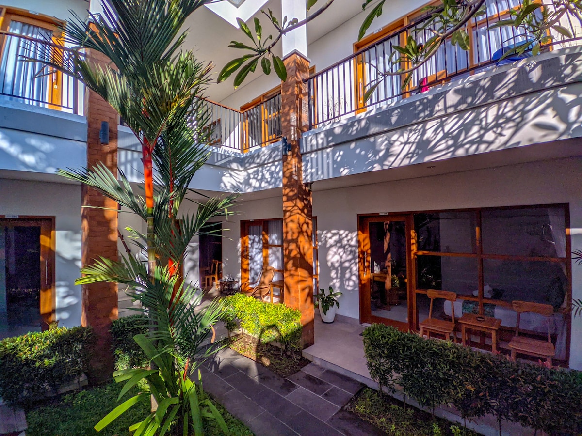 A serene villa courtyard is framed by lush greenery and tropical plants. Sunlight filters through the leaves, casting playful shadows on the walls of the open rooms. Each room features a private entrance with outdoor seating arrangements visible, enhancing the inviting atmosphere.