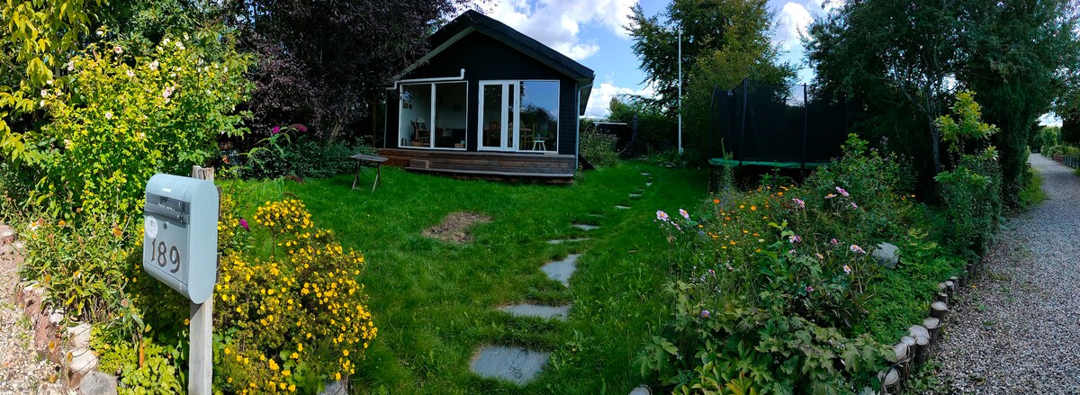 A modern house with large glass doors opens to a grassy garden, featuring pathways lined with stones and vibrant flower beds. The lush greenery surrounds the home, complemented by trees and a small outdoor seating area. The setting is peaceful and inviting for outdoor relaxation.