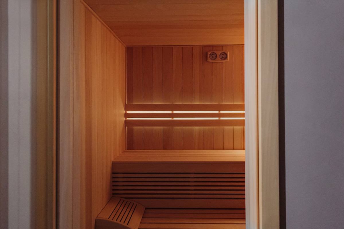 An indoor sauna features warm wood paneling that envelops the space. Two horizontal light strips add a soft glow, highlighting the simple wooden benches along the walls. The serene environment promotes relaxation and comfort for guests.