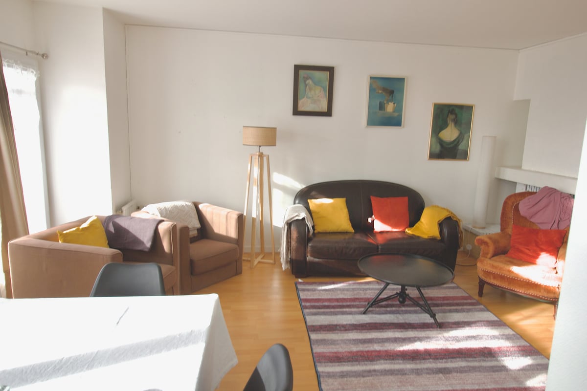 The spacious living area presents a mix of comfortable seating arrangements, including a brown leather sofa and two armchairs. A coffee table occupies the center, and artwork adorns the walls. Natural light enters through a nearby window, enhancing the inviting atmosphere.