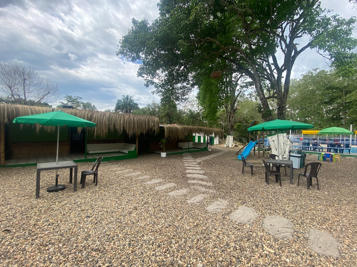 A shaded outdoor area features several thatched-roof structures arranged around a gravel pathway. Green umbrellas provide shade over black tables and chairs, with a colorful swimming area visible in the background, surrounded by lush trees.