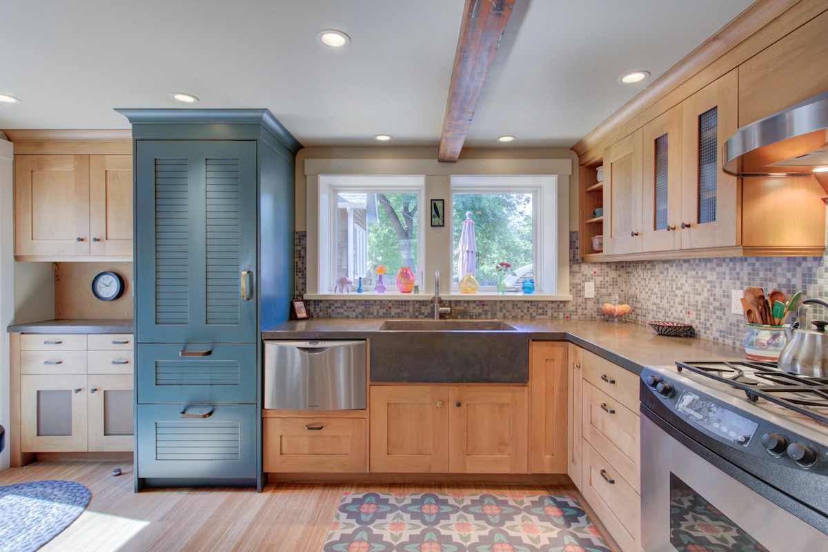 The kitchen features wooden cabinetry and a blue accent cabinet. A stainless steel refrigerator and stove are visible, along with a dark countertop and a mosaic backsplash. Natural light floods the space through a window, enhancing the warm tones of the wood.