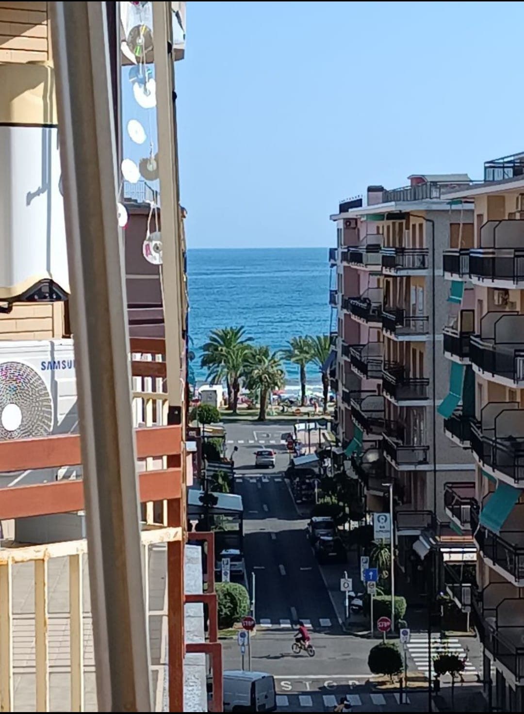 A view from a balcony reveals a street leading to the sea, with palm trees lining the waterfront. The blue ocean is visible in the background, framed by adjacent buildings. Clear skies enhance the bright and inviting atmosphere.