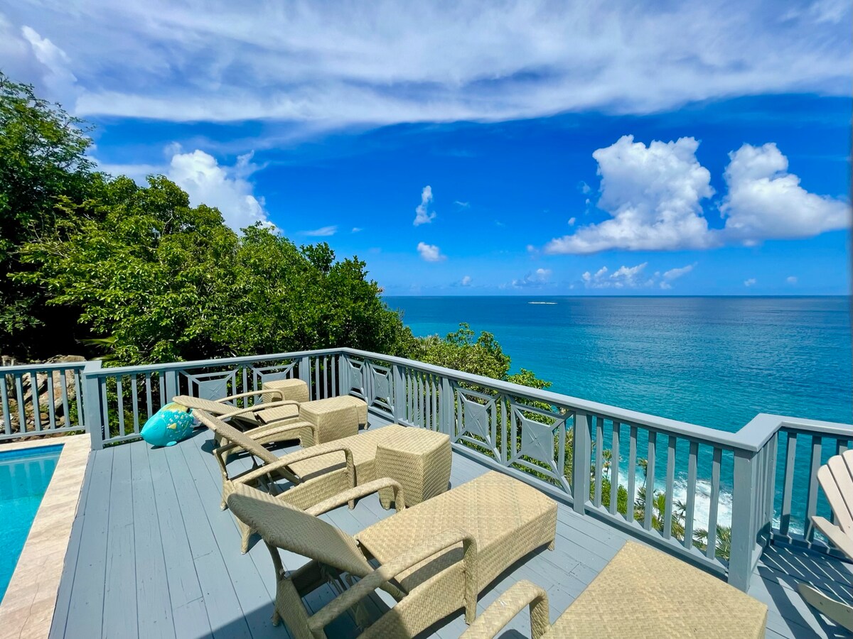A spacious deck features several lounge chairs arranged for relaxation, overlooking a panoramic view of the ocean. Lush greenery surrounds the area, and the vibrant blue sky is dotted with fluffy white clouds, enhancing the tranquil seaside atmosphere.