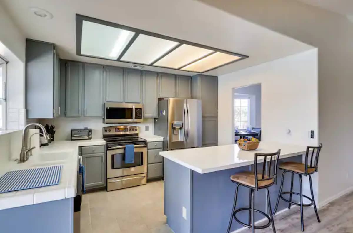 A modern kitchen features stainless steel appliances, including an oven and refrigerator. Light grey cabinets and a white countertop provide a clean and functional workspace. Two barstools are positioned at the counter, and a window allows natural light to illuminate the area.