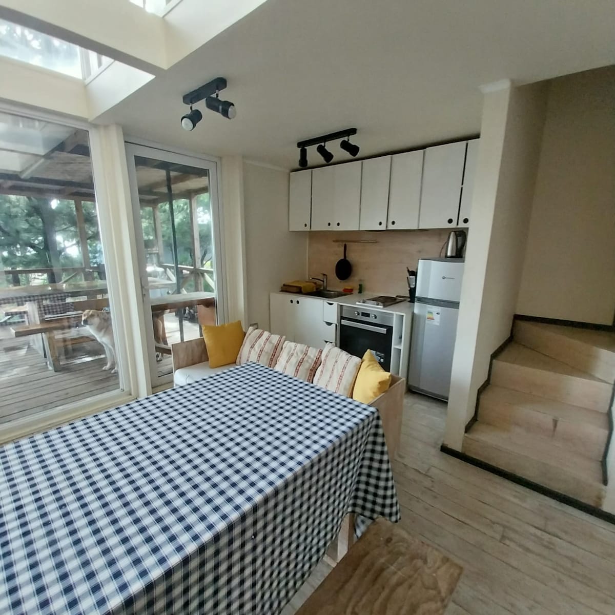 A bright dining area is highlighted by a checkered tablecloth covering a large table. Light pours in through large glass doors, while a functional kitchen setup features essential appliances. A staircase in the background leads to an upper level, enhancing the space's versatility.