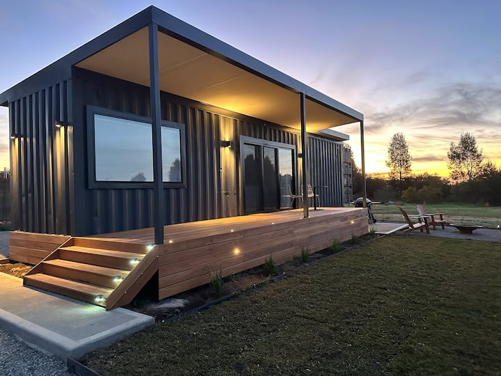 A modern tiny home made from a shipping container with a wooden deck and steps illuminated by soft lighting. The home is set against a serene sunset sky, with two outdoor chairs and a fire pit on the lawn in front of the house.