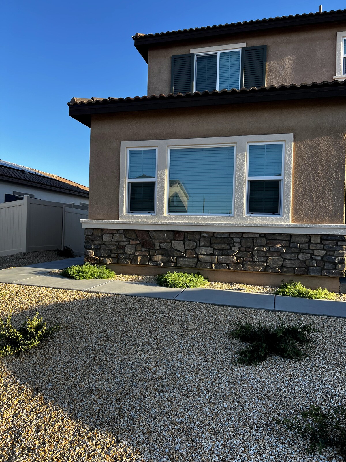 The exterior of the private guest suite is shown, featuring a combination of stone and stucco elements. Large windows allow natural light to enter, while manicured gravel landscaping complements the home's design. A clear sky serves as the backdrop, enhancing the structure's inviting appearance.