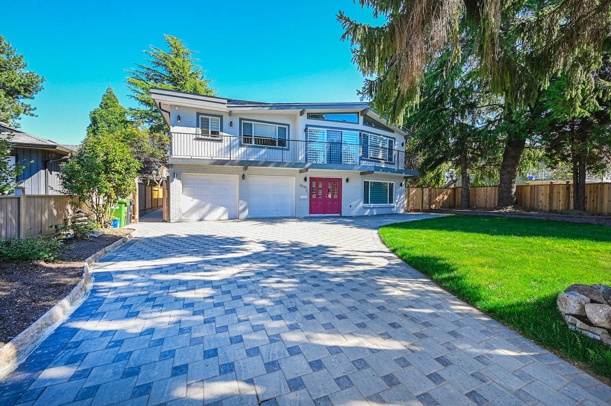 The exterior of the home features a spacious driveway made of pavers, leading to a two-story structure with a vibrant red front door and a balcony. Lush green grass and trees surround the property, creating a serene environment.