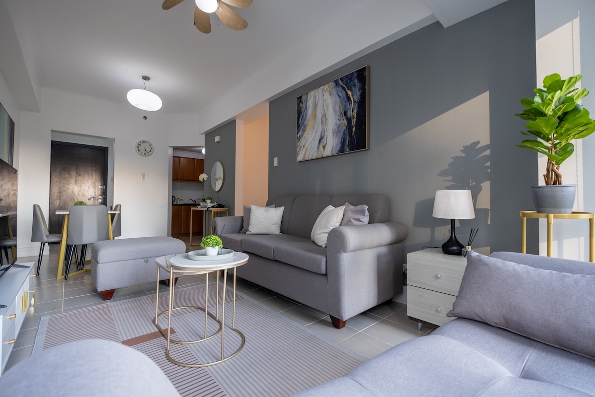 A modern living space features a pair of gray sofas flanking a circular coffee table atop a patterned area rug. A painting adorns the wall, while a plant adds a touch of greenery. Natural light enters from nearby windows, enhancing the welcoming atmosphere.