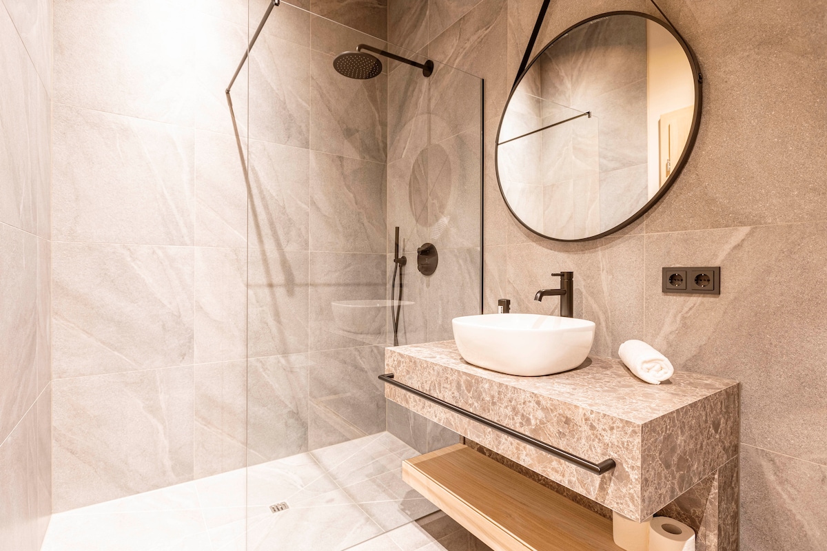 A modern bathroom features a walk-in shower with a circular rainfall showerhead and glass panel. A sleek stone vanity with a white basin is complemented by a round mirror. Open shelving displays neatly arranged towels and toiletries.