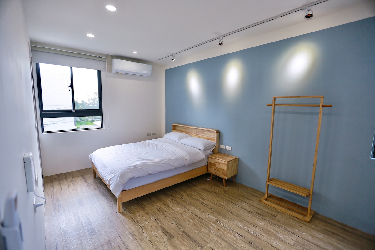 A spacious bedroom is presented with a light wood bed and bedside table. A clothing rack is positioned against the wall, and natural light enters through a large window. The soft blue accent wall complements the neutral tones, creating a calm environment.