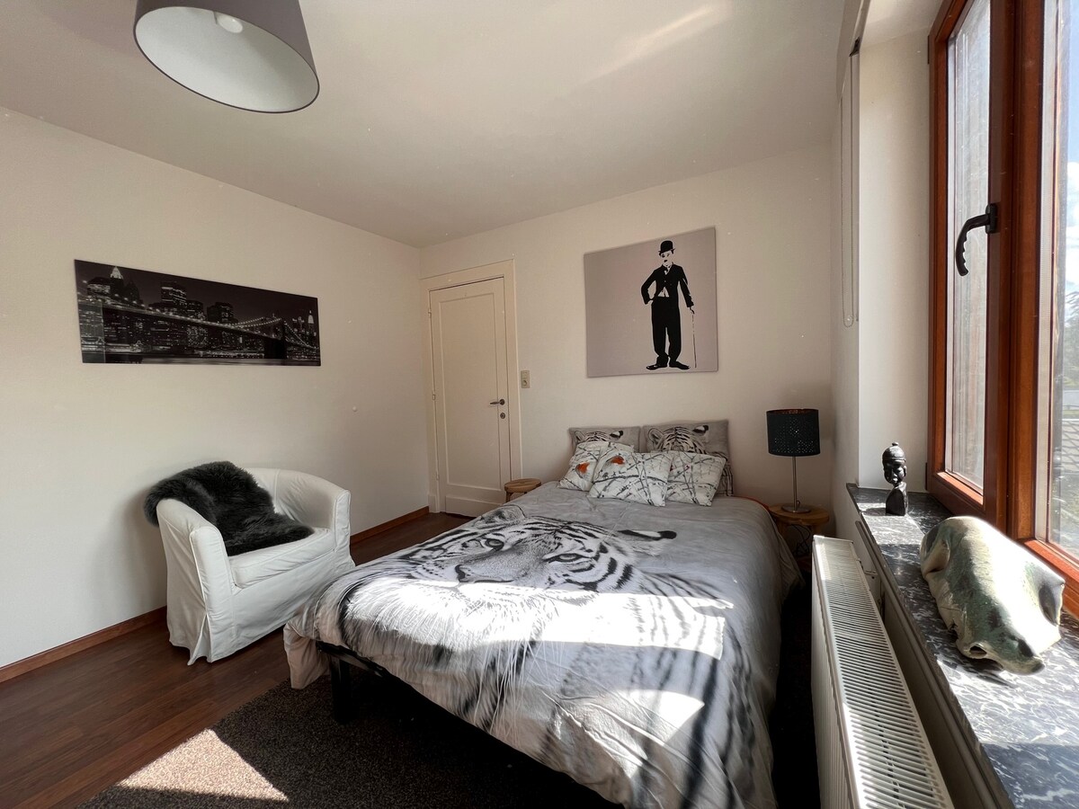 A well-lit bedroom features a spacious bed adorned with a patterned blanket. A cozy chair is positioned nearby, and a large window offers natural light. Wall art depicting cityscapes and a figure adds a contemporary touch, with additional decorative elements enhancing the room.