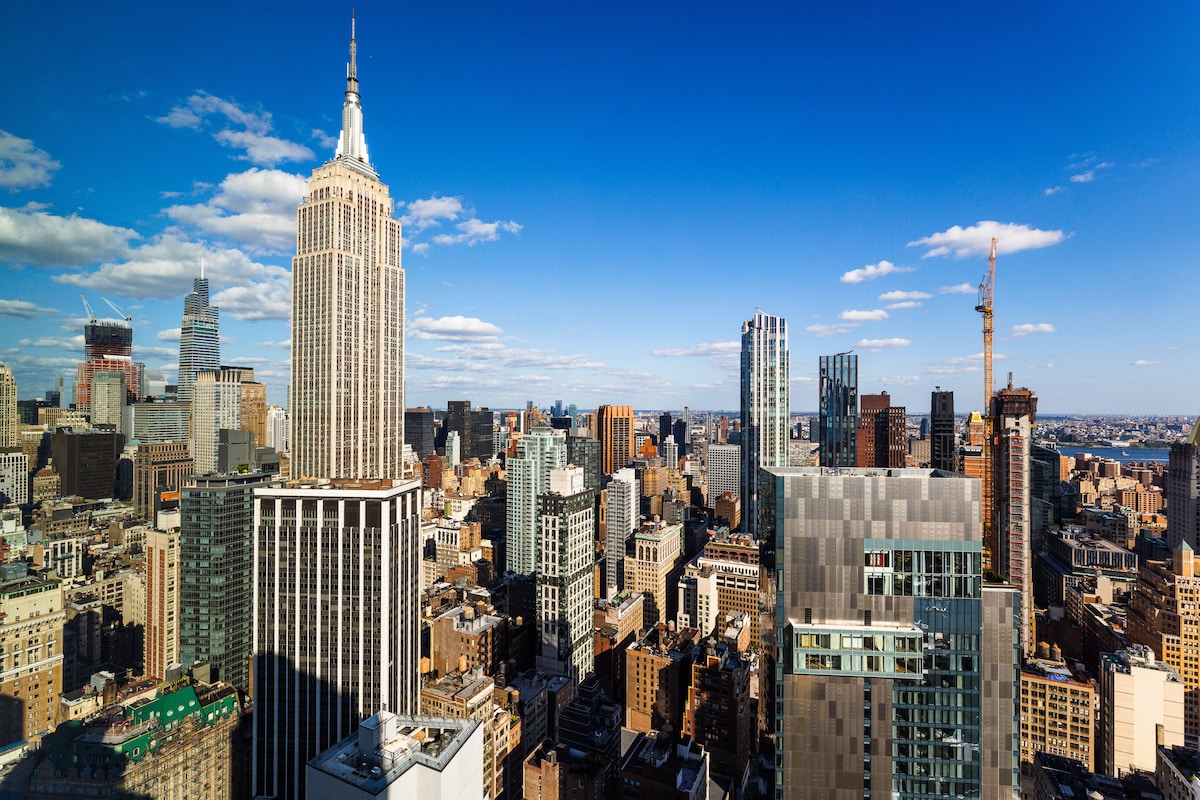 A panoramic view of the city skyline is captured, highlighting The Empire State Building prominently. Various skyscrapers fill the horizon against a clear blue sky, with the interplay of sunlight and shadows creating depth across the urban landscape.