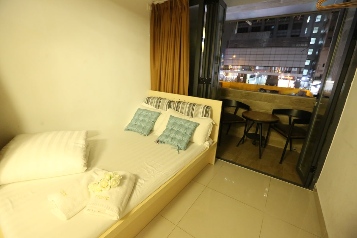 A cozy sleeping area features a spacious bed adorned with two decorative pillows. Natural light filters through the large open window, which leads to a small seating area with round tables. Warm curtains contribute to the inviting ambiance of the room.