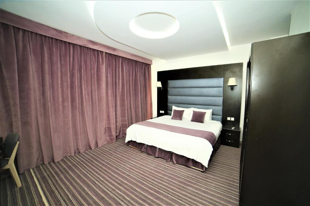 A spacious bedroom is showcased, featuring a large bed with a dark headboard and white bedding accented with purple lines. Soft purple curtains frame a wall, while bedside lamps are mounted on either side of the bed. A wardrobe is positioned nearby.