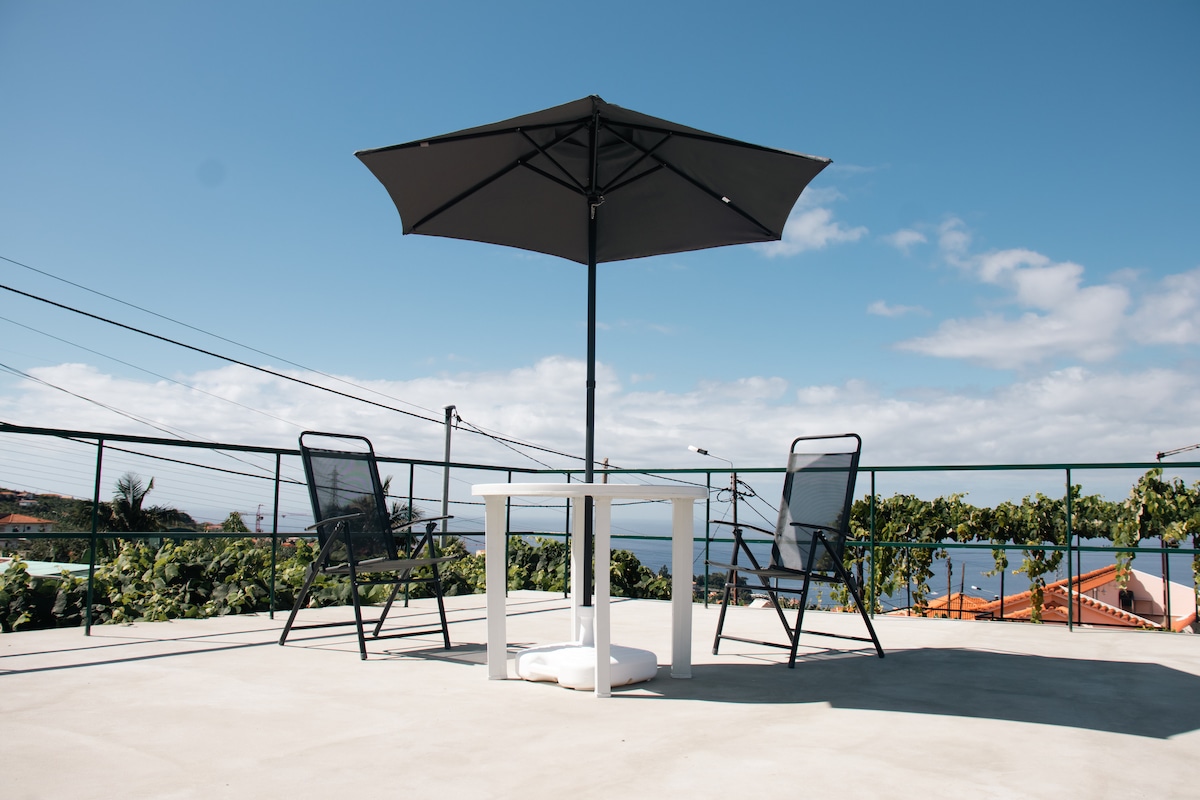 A spacious terrace features a round table with a large umbrella providing shade. Two black chairs are positioned around the table, allowing for unobstructed views of the surrounding landscape and distant ocean. The clear sky and vibrant greenery enhance the outdoor setting.