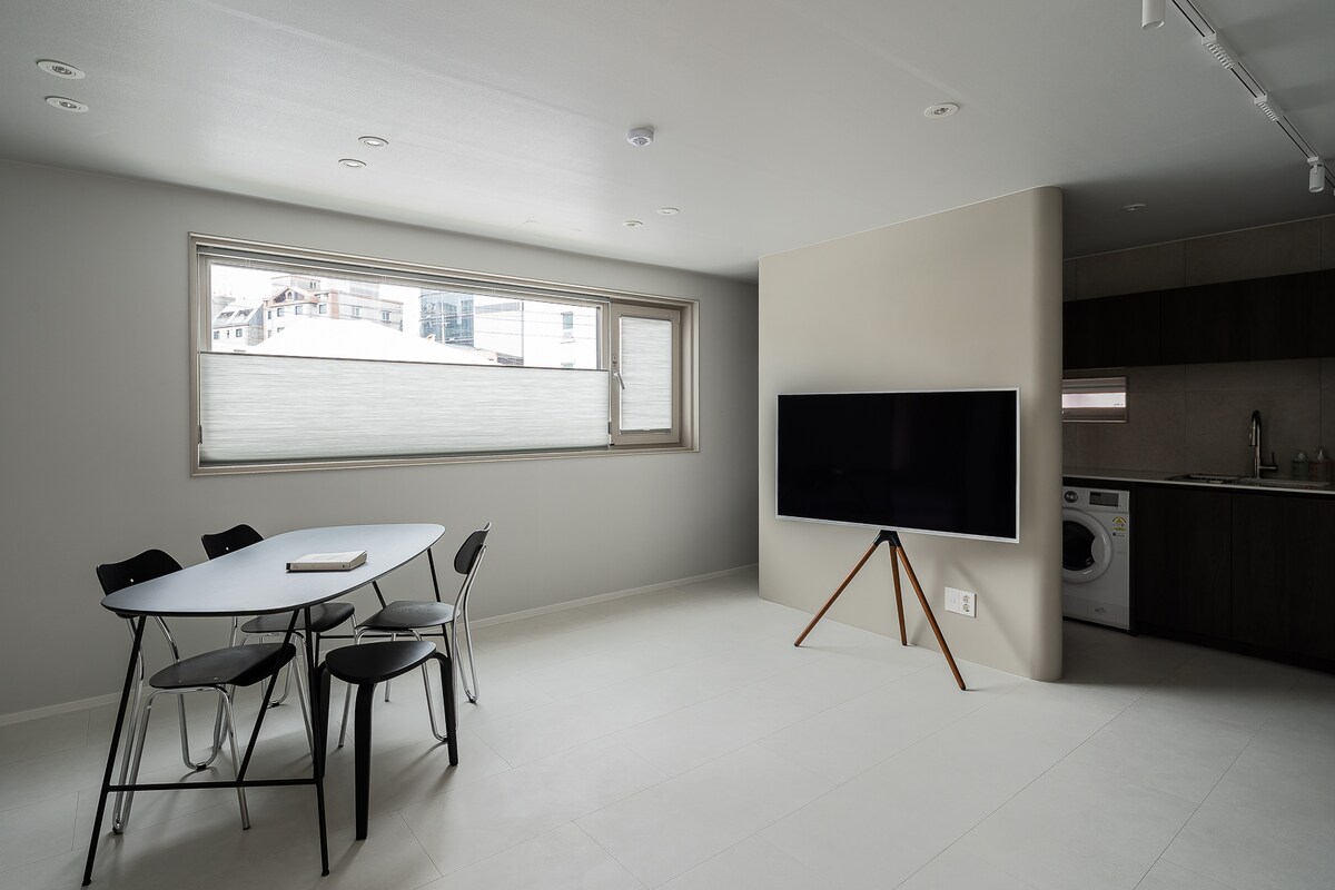 A light-filled living area features a rectangular dining table with four black chairs. A sleek television is mounted on a stand, with a washer visible in a nearby kitchen area. Large windows allow natural light to illuminate the minimalist design.