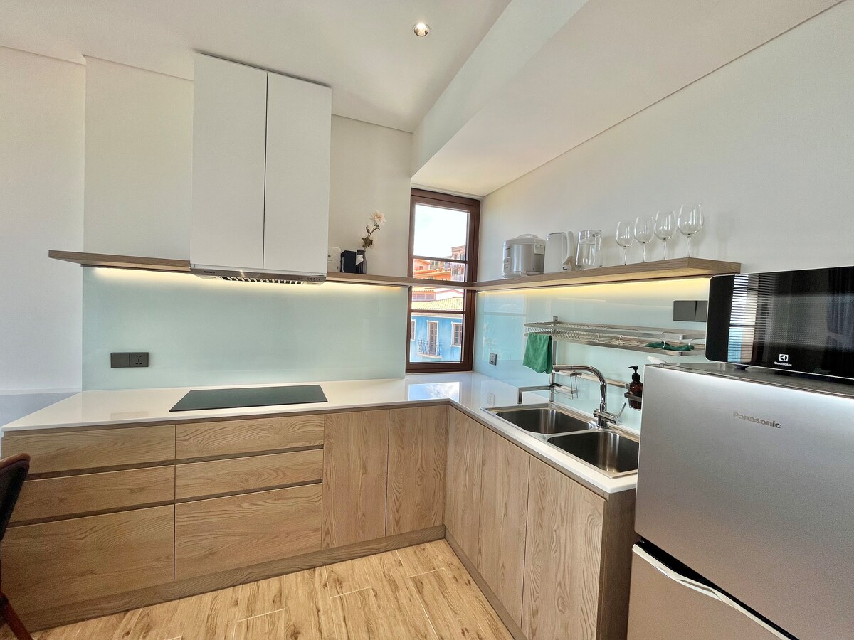 A modern kitchen features light wooden cabinetry and a well-lit countertop. Essential appliances include a refrigerator, microwave, and a stainless-steel sink with a tiled backsplash. Glassware is neatly arranged on a shelf above the counter, with a small window providing natural light.