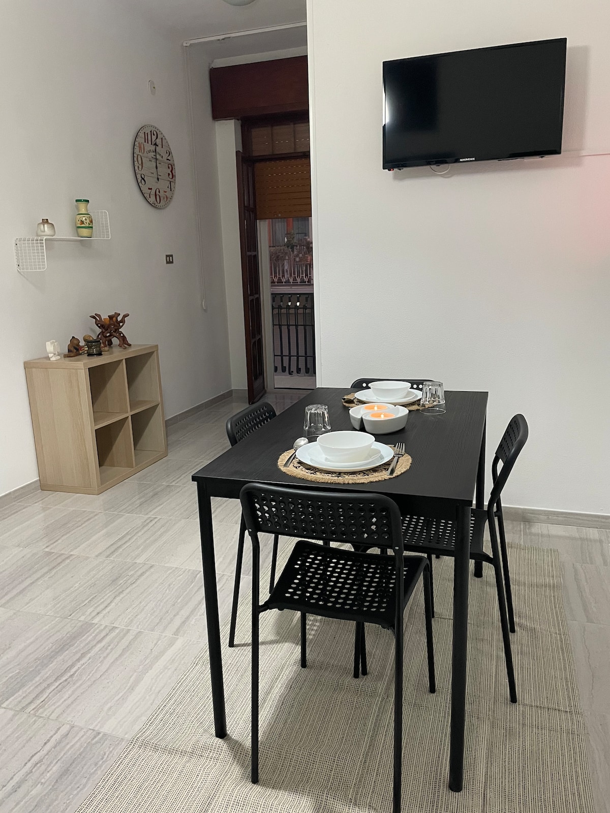 A dining area features a black table set for four, complete with dishes and glassware. Black chairs surround the table. A wall-mounted television is visible, along with a minimalist shelf and a clock on the wall, adding functionality to the space.