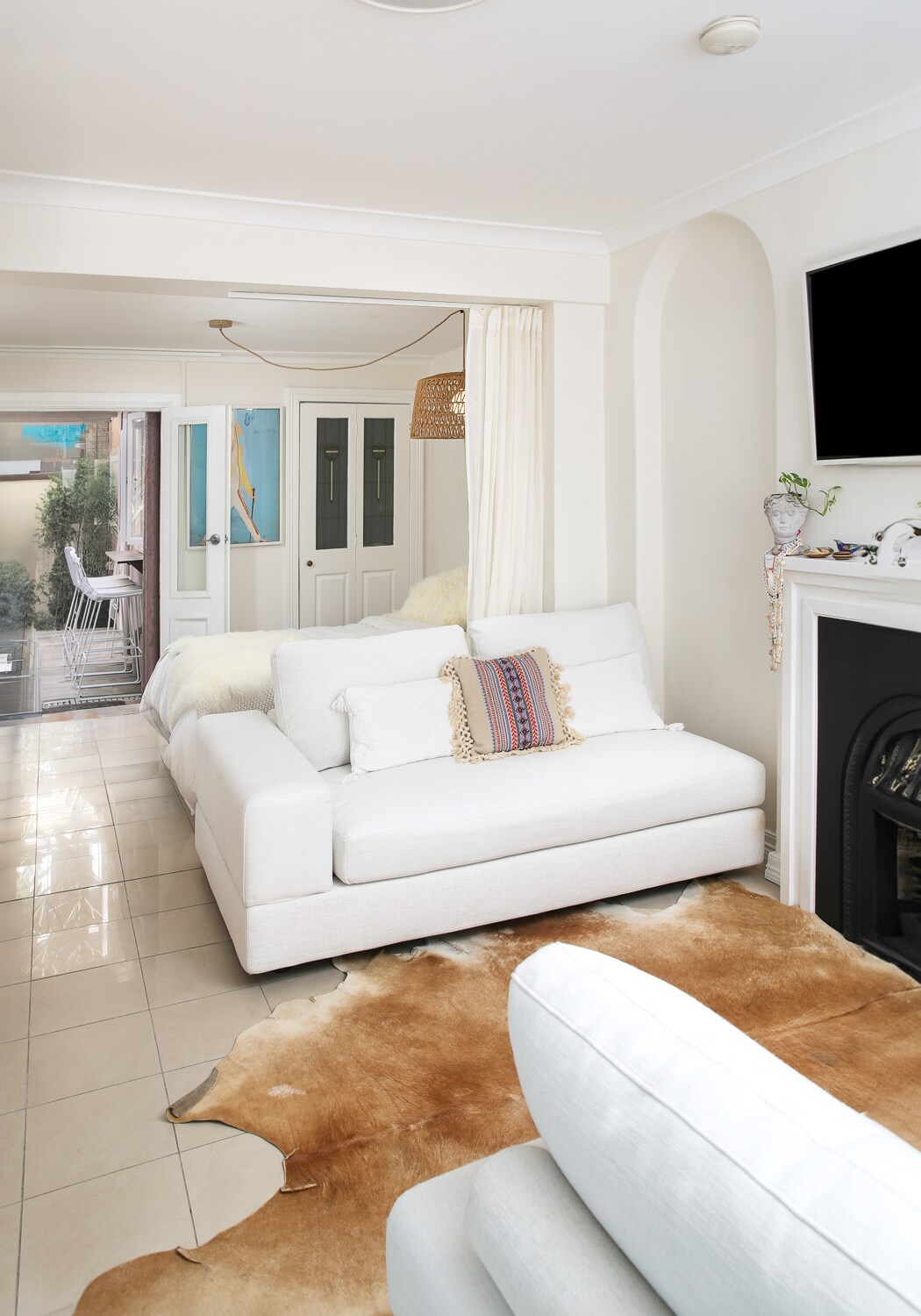A comfortable living area features a sectional white sofa adorned with a decorative pillow. A light-colored cowhide rug lays on the tiled floor, while a minimalist fireplace adds warmth. Background doors lead to a cozy outdoor space, enhancing the inviting setting.