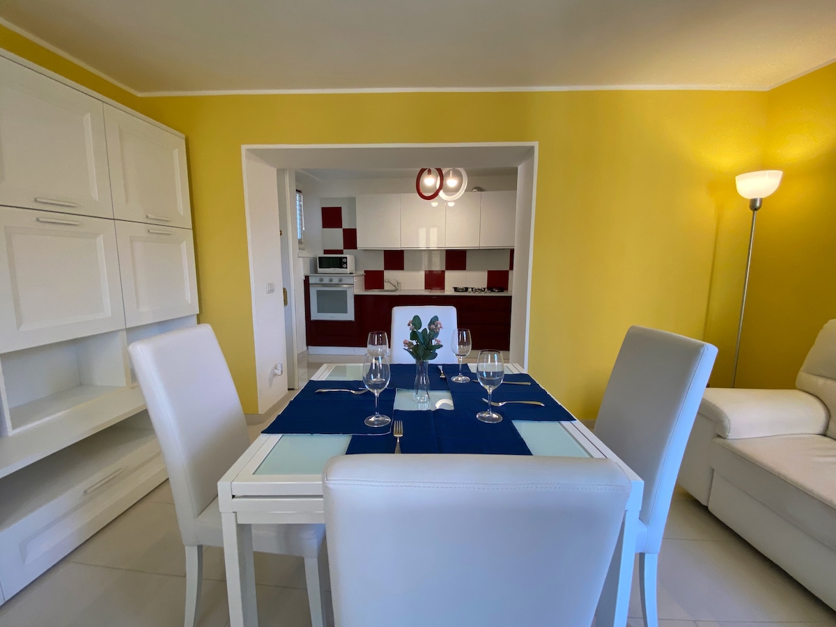 A modern dining area features a white table set with glasses and a blue table runner, surrounded by four white upholstered chairs. The vibrant yellow walls enhance the inviting atmosphere, while a lamp provides soft illumination. A glimpse of the kitchen can be seen in the background.