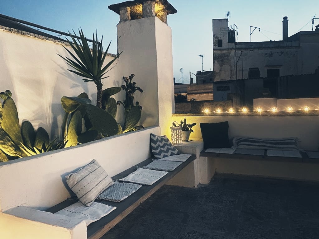 A private terrace is decorated with potted plants and soft lighting. Comfortable seating areas are created with built-in benches, featuring soft cushions. The surrounding rooftops of the historic town are visible in the background, providing a serene atmosphere for relaxation.