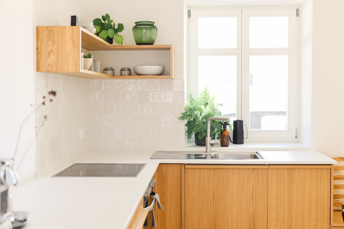 A modern kitchen features a stainless steel sink and a countertop made of light-colored material. Wooden cabinetry provides storage, while open shelves display potted plants and dishware. A window allows natural light to brighten the space, with greenery visible outside.