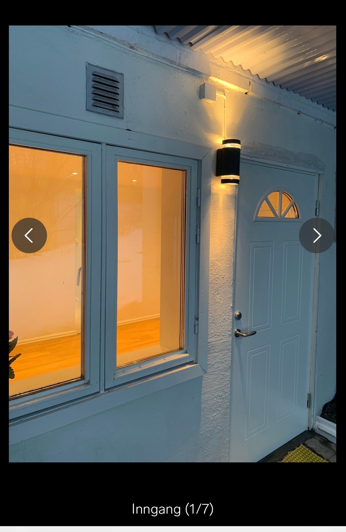 An entrance is displayed with a white door, featuring a small window at the top. Two large windows on the side are illuminated with warm light, adding a welcoming touch. A small light fixture is mounted above the door, enhancing visibility.