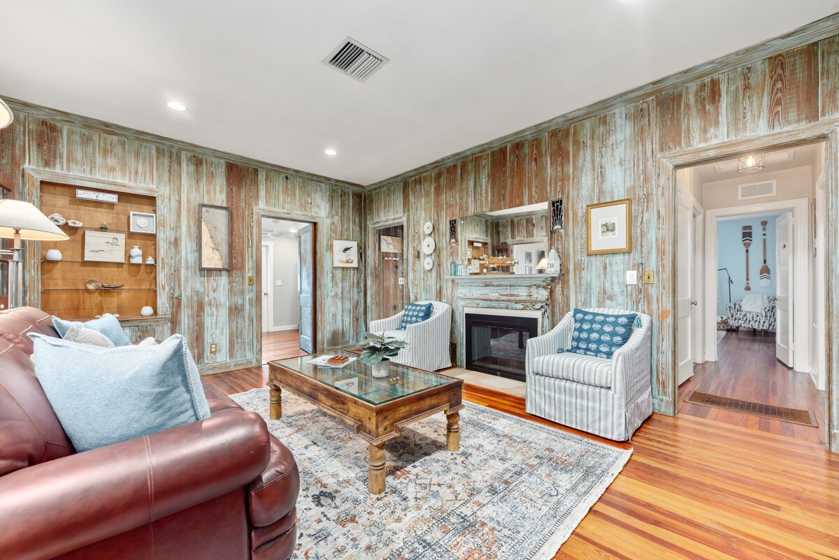 A welcoming living room features a rustic wood-paneled wall, accented by a stylish area rug and a glass coffee table. Two upholstered chairs are positioned around the table, while a cozy sofa offers additional seating. A decorative mirror is mounted above a warm fireplace.