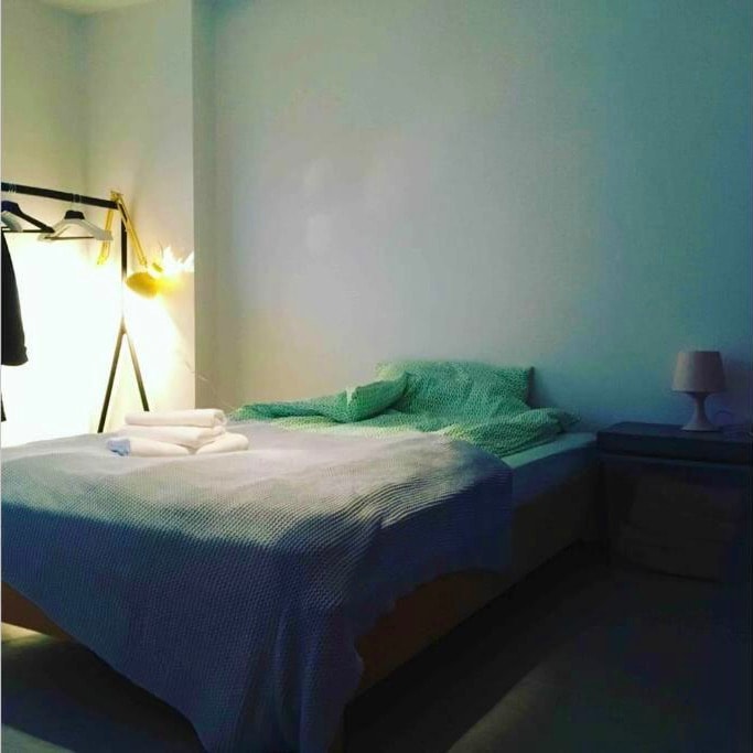 A sleeping area features a spacious bed with a light green patterned blanket and pillows. A bedside table with a lamp is positioned next to the bed, and a clothing rack stands against the wall, illuminated by a warm light.