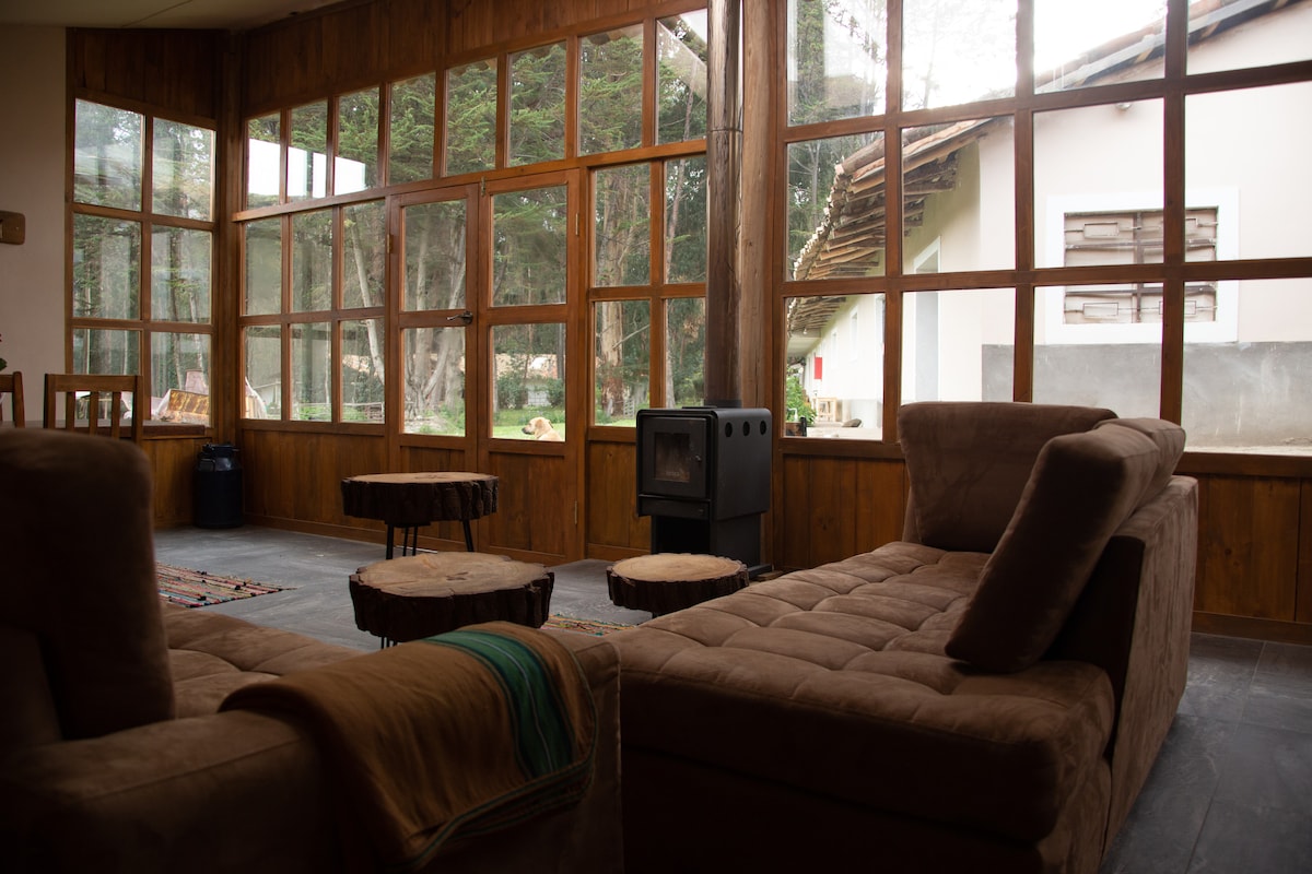 A spacious lounge area is featured with large windows that invite natural light. Comfortable lounge sofas are arranged around wooden coffee tables, creating a cozy space. A small wood stove is placed against a wall, adding warmth to the inviting environment.