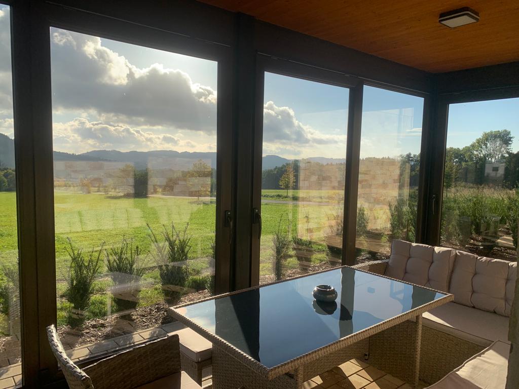 A sunlit sunroom features large glass panels that offer views of the surrounding greenery and distant hills. A modern glass-topped table is surrounded by comfortable seating, creating an inviting space for relaxation and gatherings.