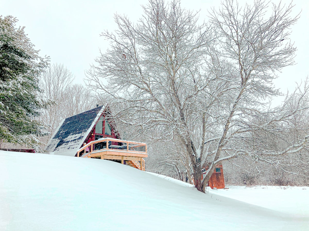 A cozy A-frame cabin is nestled in a snowy landscape, surrounded by a blanket of fresh, white snow. A large tree stands nearby, partially covered in snow, with a small shed visible in the background. The warm wood deck invites relaxation amid the winter scenery.