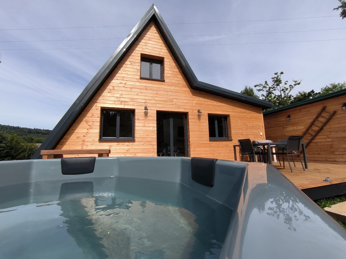 A wooden A-frame house is displayed, featuring large windows that allow natural light to illuminate the interior. In the foreground, a private hot tub invites relaxation, while the spacious terrace showcases a dining area with chairs. Lush greenery surrounds the property.