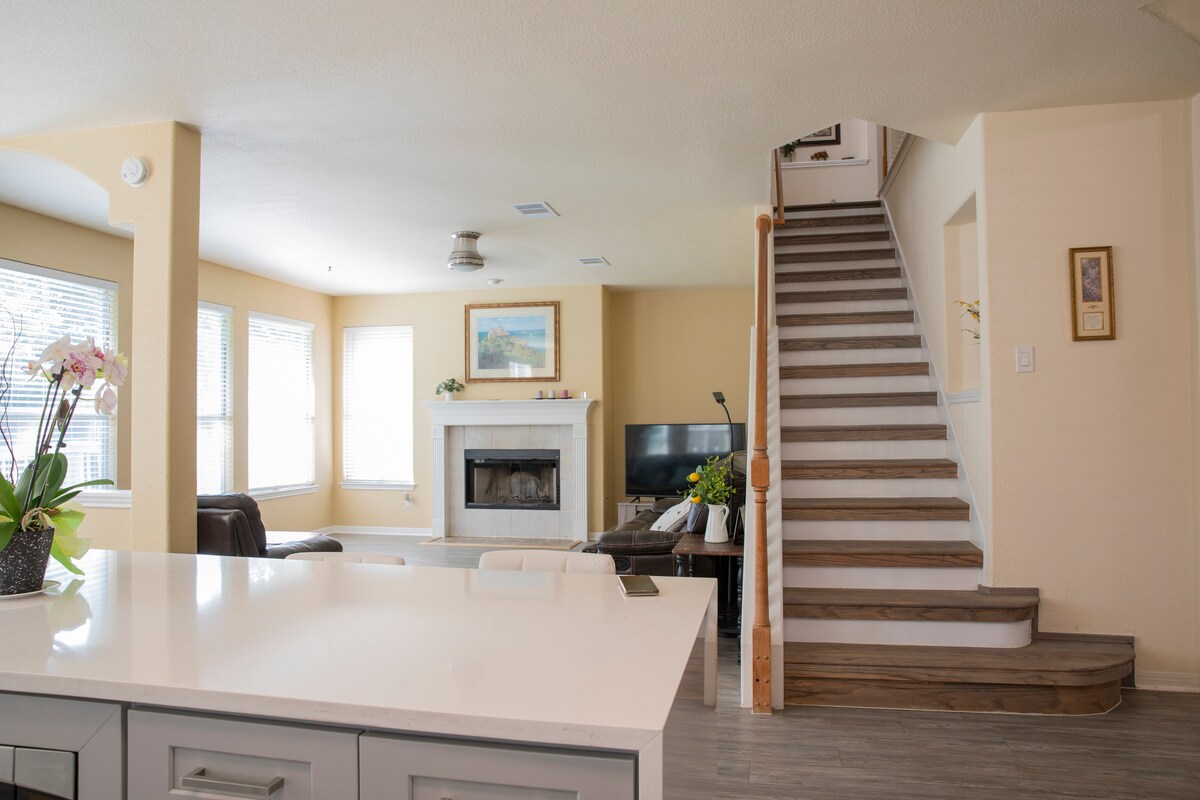 An open living area features large windows allowing natural light to fill the space. A fireplace is situated between built-in shelving. A modern kitchen island is positioned in the foreground, while a staircase leading to the upper floor is visible in the background.