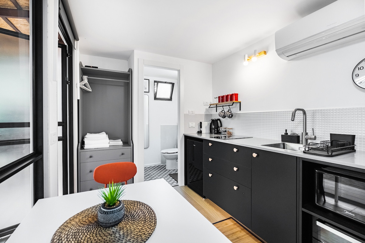 A modern kitchen area is presented with sleek black cabinetry and a white countertop. The space includes a small dining table with an orange chair, and essential appliances are visible. A closet and bathroom door are noted in the background, contributing to the overall functionality.